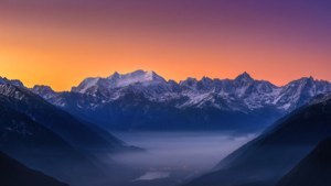 सूर्योदय के समय बर्फ से ढके पहाड़ों का लुभावना अल्ट्रा-वाइड दृश्य, जिसमें धुंध भरी घाटी है।
